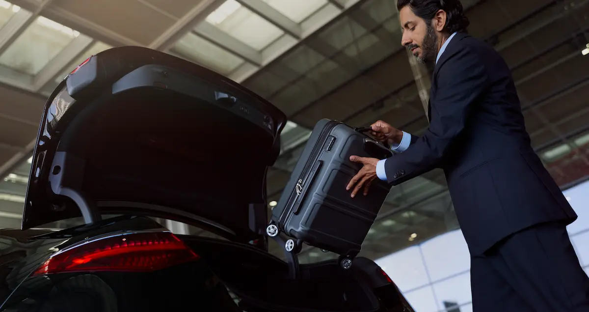 A Blacklane chauffeur places a luggage case in the trunk of a black Mercedes limousine outside an airport terminal.