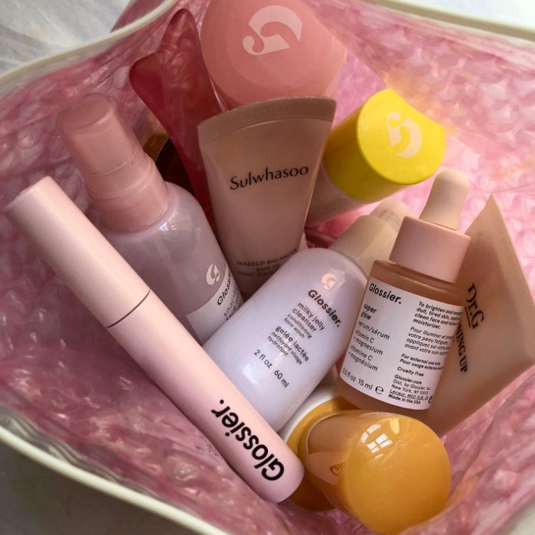 Lash Slick, Soothing Face Mist, Milky Jelly Cleanser, Solution, Invisible Shield, and Super Glow with other makeup products in a Glossier pink pouch.