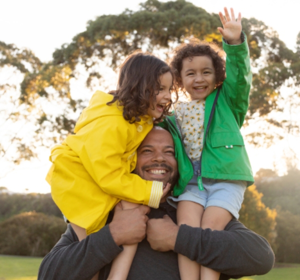 A father stands smiling whilst lifting his two kids onto his shoulders.