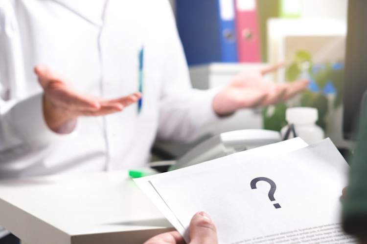 person holds sheet of paper with question mark on it as another person shrugs in background
