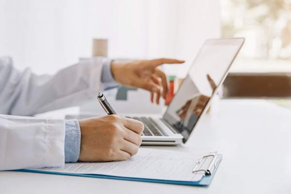Doctor writing medical documentation and using laptop