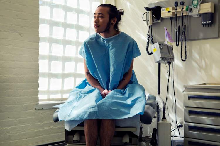 A transgender person sitting in a hospital gown sitting in an exam room