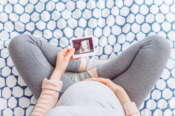 pregnant young woman holding hand on her belly, sitting on bed and hold ultrasound image of baby