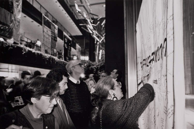 Archival photo of concertgoers writing the names of loved ones on a special panel of the AIDS Memorial Quilt in 1992