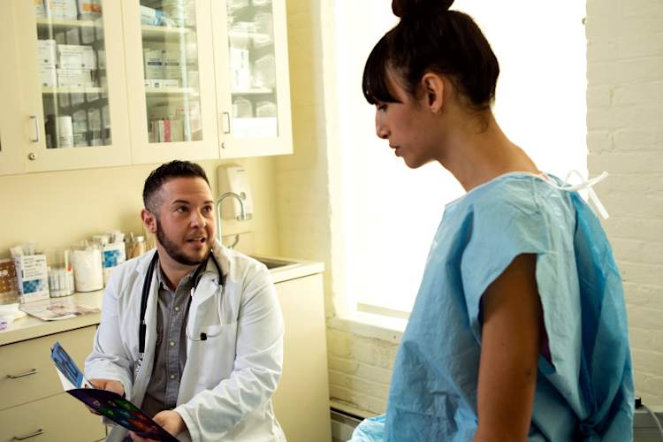A transgender woman in a hospital gown speaking to her doctor, a transgender man, in an exam room