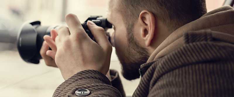 This is a photo of a male in a brown jacket. The man is taking a photograph. He appears to be spying on someone or trying to take the photograph discreetly.