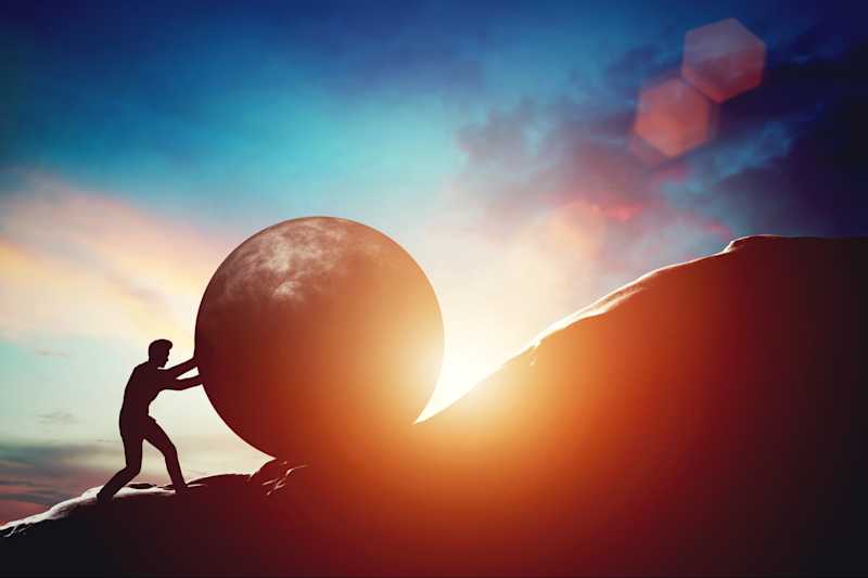 A photo of a human being pushing a large round rock up the mountain.