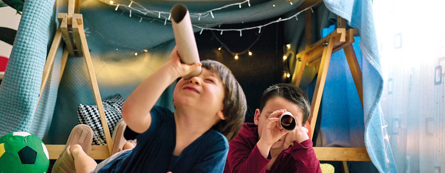 Two children play in the living room in a tent built with tables and blankets
