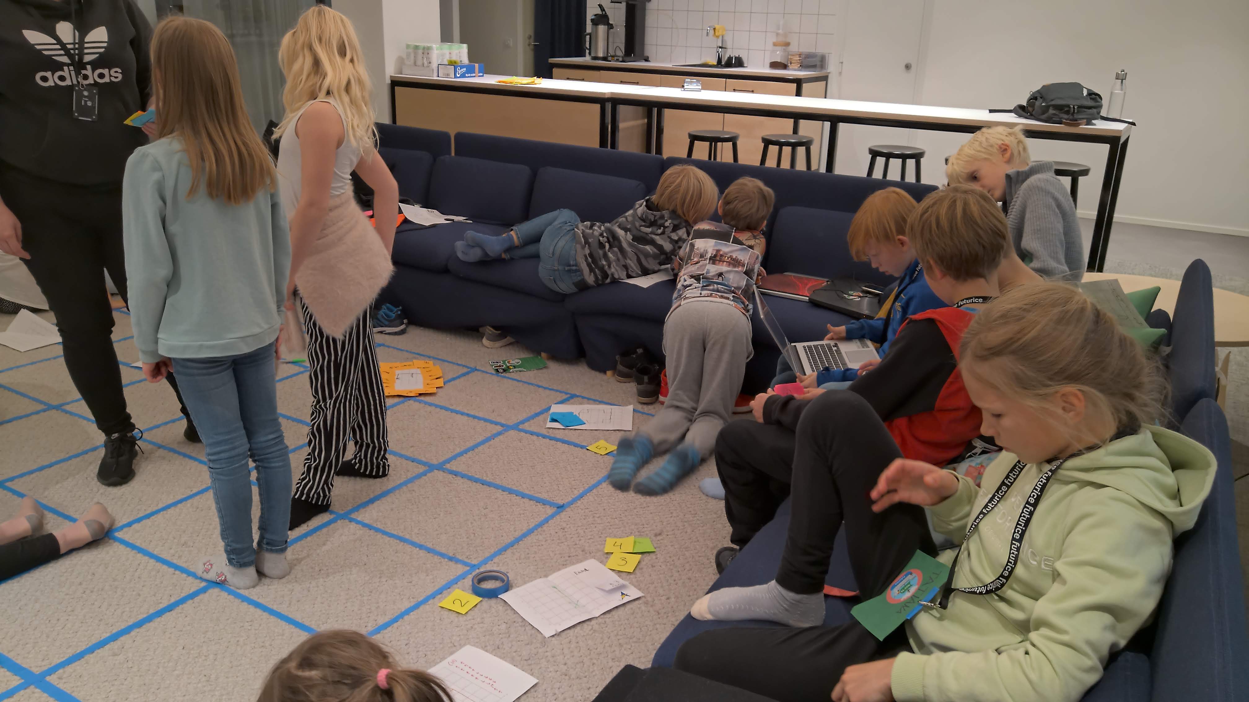 A square grid on the floor made with blue tape, children playing around the floor and sofas