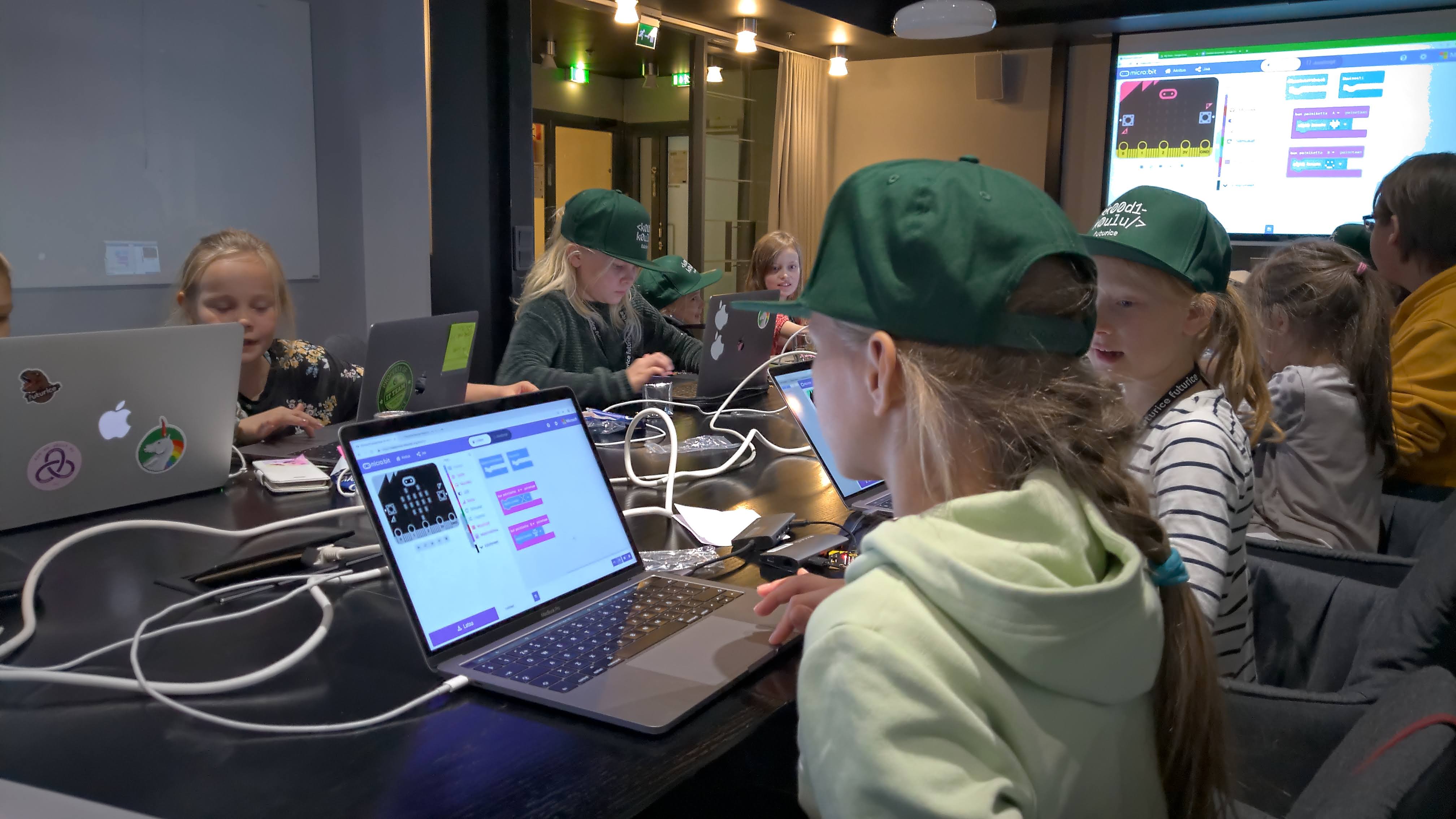 A group of children with green caps sitting in a meeting room working with laptops