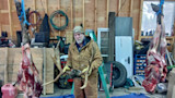 Man in workshop holding large antlers between two hanging skinned carcasses, ladder labeled KELLER behind him