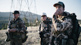 Three hunters in camouflage gear by a transmission tower; center smiles, right covers mouth