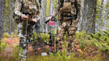 Two hunters in camouflage stand on mossy forest floor holding compound bows with pink-fletched arrows