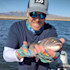 Man on boat holding trout toward camera, wearing gray cap, mirrored sunglasses, colorful fingerless gloves