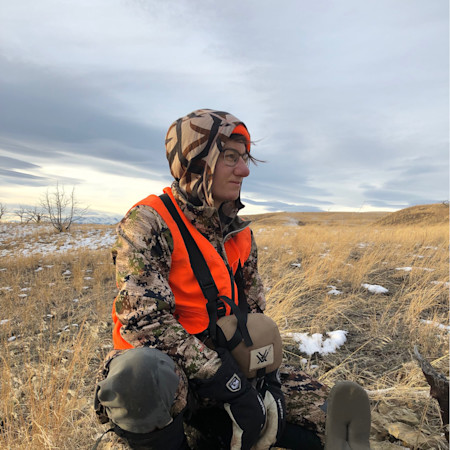 Hunter kneeling in grassy field wearing camouflage, orange safety vest, hood and glasses