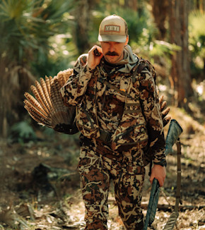 Hunter in camouflage carrying a turkey and shotgun, wearing cap reading "YETI"