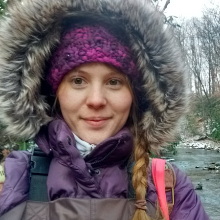 Woman in fur-lined hood and purple jacket, purple knit headband, braid by a stream