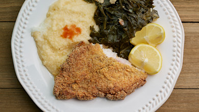 Fried catfish with grits, collard greens, and lemon slices on white plate