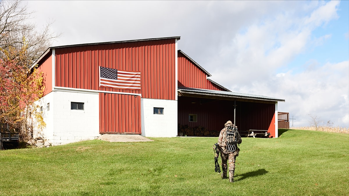 Hunter in camouflage carrying a bow walking toward a red barn with an American flag
