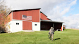 Hunter in camouflage carrying a bow walking toward a red barn with an American flag