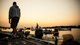 Man standing on boat at sunrise with multiple fishing boats and a bridge on the horizon
