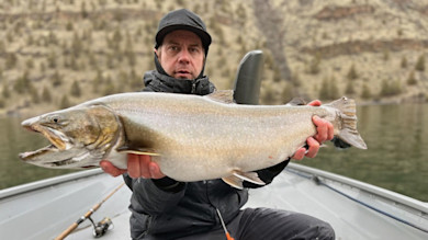 Man holding large bull trout in a boat on a lake with rocky shoreline behind