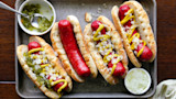 Five venison hot dogs in buns on baking tray with mustard, diced onions, and relish