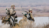 Two hunters in camouflage carrying dozens of duck decoys and a shotgun along a lakeshore