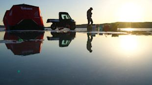 Angler silhouetted ice fishing at sunrise with red Eskimo shelter, UTV, and reflections