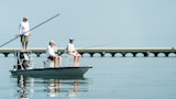 Three men fishing on a skiff near an arched bridge; one stands on poling platform; boat labeled FL 0902 PX