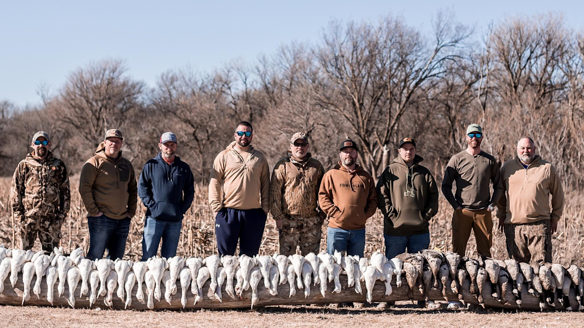Nine hunters standing behind a log displaying dozens of white snow geese in a field
