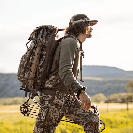 Hunter in camouflage carrying a large pack and compound bow in grassy field