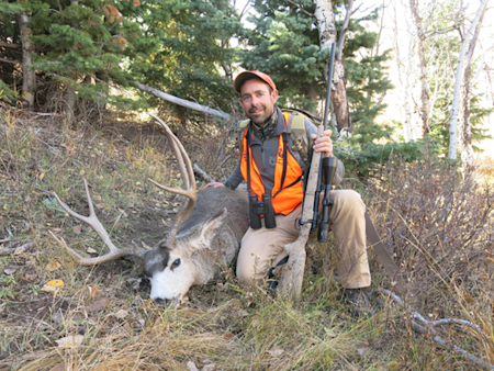 Hunter kneeling beside large mule deer buck, holding rifle, wearing orange cap and safety vest