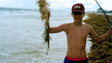 Shirtless boy on beach holding catfish by seaweed, fishing rod visible, wearing cap reading "FSU"