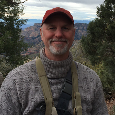 Smiling man in red cap and gray knit sweater wearing a binocular harness with canyon background