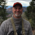 Smiling man in red cap and gray knit sweater wearing a binocular harness with canyon background