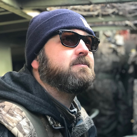 Bearded man wearing sunglasses and knit beanie, camo jacket, looking upward