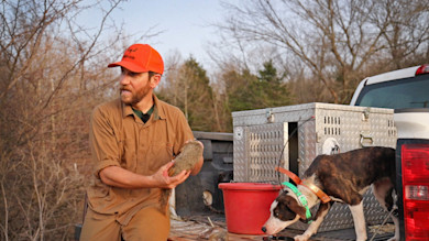 Hunter holding skinned squirrel on pickup tailgate, dog beside metal dog box labeled MEATEATER