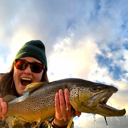 Woman holding large brown trout, wearing green beanie and sunglasses