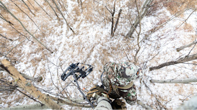 Hunter in camouflage standing on a tree stand holding a compound bow; overhead view of snowy forest floor