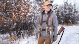 Man in gray jacket wearing orange cap labeled First Lite, holding rifle and two squirrels in snowy field