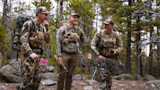 Three hunters in camouflage baselayers and backpacks holding compound bows in pine forest