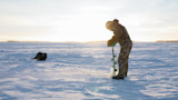 Man drilling hole with green ice auger on frozen lake at sunrise, black pack on snow