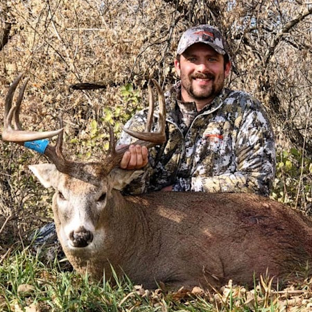 Smiling hunter kneels behind a harvested whitetail buck, holding its antlers.
