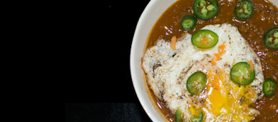 White bowl of chili topped with a fried egg and sliced jalapeños