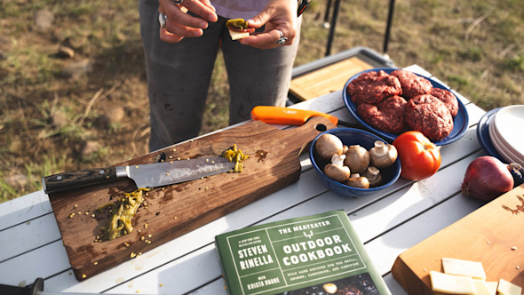 Camp prep: knife on cutting board, mushrooms, burger patties, tomato, and book THE MEATEATER OUTDOOR COOKBOOK