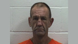Mugshot of middle-aged man in orange jail shirt standing against cinderblock wall