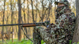 Hunter in full camouflage and face veil, seated against a tree aiming a double-barrel shotgun in woods