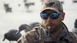 Hunter blowing a duck call wearing mirrored sunglasses and camo cap; decoy ducks floating behind