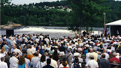 Crowd gathered on riverbank facing a low concrete dam, speakers at podium under tent and American flag